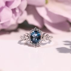 an oval blue stone surrounded by diamonds sits on a white surface next to pink flowers