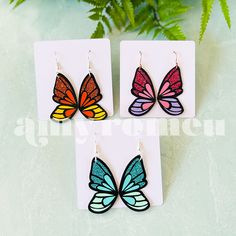 three pairs of butterfly shaped earrings on top of a white card with green plant in the background