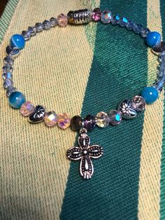 This silver-pated cross charm bracelet is filled with little surprises, like silver metal heart beads, light pink aurora borealis faceted glass beads, striped blue agate, and light blue glass beads. This bracelet is sized for a child. Silver Crystal Bracelet With Heart And Round Beads, Silver Crystal Bracelet With Heart Shaped Beads, Silver Spiritual Bracelet With Heart Beads, Silver Beaded Spiritual Charm Bracelet, Adjustable Silver Beaded Bracelets With Cross Shape, Blue Hypoallergenic Charm Bracelet With Round Beads, Silver Spiritual Beaded Charm Bracelet, Adjustable Silver Cross Beaded Bracelets, Adjustable Blue Rosary Bracelet With Cross