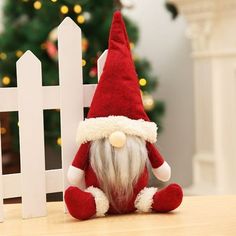 a red and white santa claus doll sitting in front of a fence next to a christmas tree