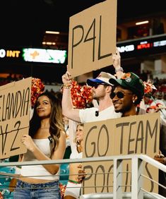 two people holding placares in the stands at a football game, one is wearing a hat and the other has a sign that says go team