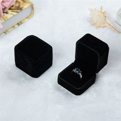 an open black velvet ring box sitting on top of a white surface next to a pink flower