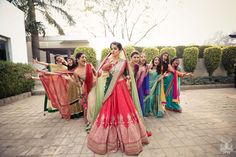 a group of women dressed in colorful sari dancing on a brick walkway with trees and bushes behind them