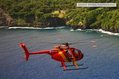 a red helicopter flying over the ocean next to a lush green forest covered shore line