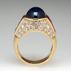 This exquisite ring is centered with an oval, sapphire cabochon in a partial bezel setting. The shoulders are each channel set with four (4) square mixed cut rubies. The top of the ring is bead set with a total of forty-six (46) round brilliant cut diamonds. The ring measures 13.7mm at the top, rises 11.2mm above the finger, tapering to 5.5mm wide and 1.3mm thick at the base of the shank. This ring is currently a size 7. Elegant Cabochon Sapphire Ring For Formal Occasions, Luxury Sapphire Cabochon Ring For Anniversary, Elegant Formal Cabochon Sapphire Ring, Elegant Formal Sapphire Cabochon Ring, Luxury Domed Cabochons For Formal Occasions, Luxury Domed Cabochons For Anniversary, Luxury Cabochon Sapphire Ring For Wedding, Elegant Cabochon Sapphire Ring, Formal Domed Cabochon Sapphire Ring