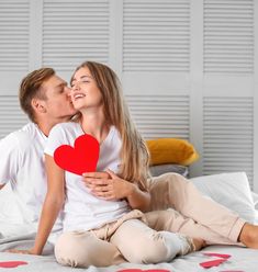 a man and woman sitting on top of a bed kissing with a heart shaped pillow