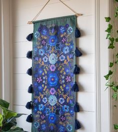 a blue wall hanging with tassels and flowers on it next to a potted plant