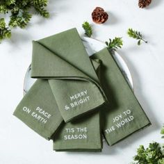 green napkins with merry and bright words on them sitting on a white plate surrounded by greenery