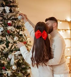 a man and woman standing next to a christmas tree with red bows on their heads