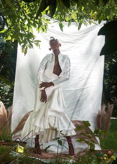 a woman in a white dress standing next to a large piece of cloth hanging from a tree