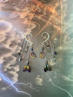 This enchanting mermaid moon ear cuff is sure to make a statement and bring out your inner magic! Silver ear cuff is embellished with a charm of two women in the moon, rainbow mermaid tail charm, iridescent drop crystals, seashell and starfish, and electroplated lava stone beads all dangling from silver and rainbow chains. This ear cuff is perfect for adorning pierced and non-pierced ears, and is lightweight and comfortable to wear. Lava Stone offers the following positive energies for the weare Fantasy Style Festival Ear Cuff, Fantasy Style Festival Ear Cuff For Pierced Ears, Handmade Fantasy Metal Cartilage Earrings, Handmade Metal Fantasy Cartilage Earrings, Bohemian Metal Dangle Body Jewelry, Fantasy Style Silver Ear Cuff For Festivals, Unique Ear Cuff With Ear Wire For Festivals, Unique Single Ear Cuff For Festivals, Whimsical Dangle Jewelry For Festivals