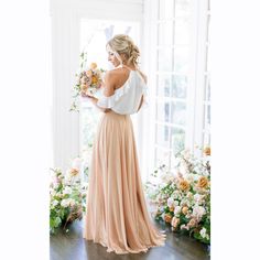 a woman standing in front of a window with flowers on the floor and wearing a long skirt