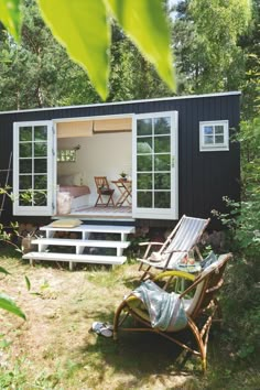 a small black cabin in the woods with two lawn chairs and an open patio door