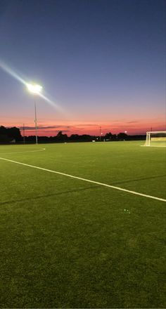 an empty soccer field at night with the sun setting in the distance and lights on