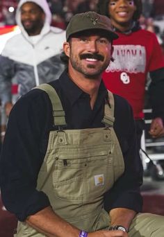 a man in overalls and a hat is smiling at the camera with other men behind him