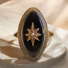 Victorian Nicolo Onyx Ring A stunning relic of the Victorian era, this Nicolo onyx ring is a masterpiece of elegance and symbolism. Its elongated marquise-shaped face is crafted from deep, jet-black onyx, a material often revered for its protective and grounding properties. At the center, a radiating star motif, meticulously rendered in warm gold, holds a single luminous pearl, symbolizing purity and celestial guidance. The detailed gold frame surrounding the onyx exudes timeless craftsmanship, Vintage Onyx Ring Art Deco, Victorian Rings Vintage, Vintage Onyx Ring, Oura Ring, Black Gemstone Ring, Onyx Rings, Obsidian Ring, Unique Rings Vintage, Victorian Ring