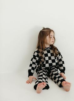 A young girl with long hair sits on the floor against a white wall, dressed in the Knit Pant Set | Black Checkerboard by forever french baby. She appears to be looking off to the side with a thoughtful expression. Black Loungewear Sets For Winter, Black Sweater For Loungewear, Black Loungewear Sets For Fall, Black Sets For Fall Loungewear, Sack Dress, French Baby, Santa Photos, Adult Pajamas, Pajama Romper