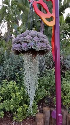 a purple pole with flowers hanging from it's sides in front of some bushes and trees