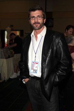 a man wearing glasses and a leather jacket standing in a room with people around him