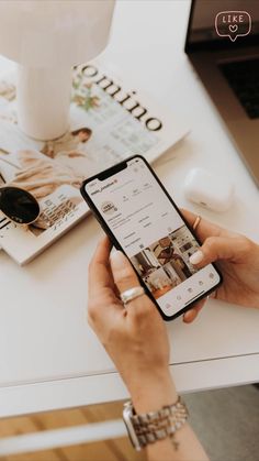 a person holding an iphone in their hand while sitting at a table with magazines and a lamp