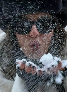 a woman wearing sunglasses and a hat is blowing snow in the air with her hands