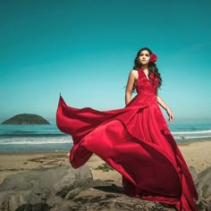 a woman in a long red dress standing on rocks near the ocean with her hair blowing in the wind