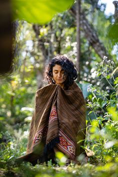 a woman wrapped up in a blanket sitting on the ground surrounded by trees and bushes