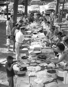 an old black and white photo of people eating food at a picnic table with cars in the background