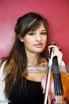 a woman with long hair holding a violin in front of a red wall and looking at the camera