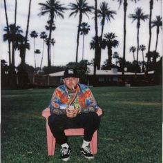 a man sitting on top of a pink chair in front of palm trees and buildings