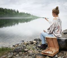 a woman sitting on top of a suitcase next to a body of water with trees in the background