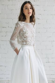 a woman standing in front of a brick wall wearing a white dress with sheer sleeves