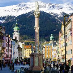 a clock tower in the middle of a town with snow covered mountains behind it and people walking around