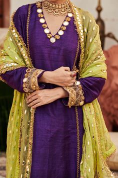 A striking ensemble, this purple raw silk sharara set showcases exquisite mirror work on the neckline and sleeves of the tunic, exuding elegance and charm. Paired with matching pants and a contrasting lime green organza dupatta, this outfit beautifully balances richness and vibrancy, making it a perfect choice for festive celebrations and special events. Anarkali Purple Palazzo Set With Gota Work, Designer Purple Chanderi Palazzo Set, Purple Anarkali Palazzo Set In Chanderi, Purple Slub Silk Wedding Set, Designer Purple Kurta With Gota Work, Purple Anarkali Chanderi Palazzo Set, Anarkali Purple Palazzo Set With Dabka Work, Purple Palazzo Set With Mirror Work For Festive Occasions, Festive Purple Palazzo Set With Dupatta