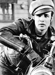 black and white photograph of a man on a motorcycle wearing a leather jacket, hat and gloves