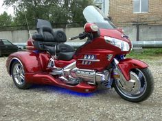 a red motorcycle parked on top of a gravel covered field next to a building and trees