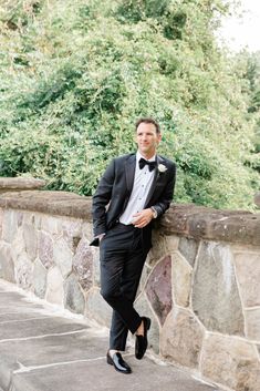 a man in a tuxedo leaning against a stone wall with trees in the background