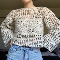 a woman standing in front of a bed wearing a white crochet sweater and jeans