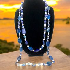 This stunning necklace measures 35 inches and features blue lapis lazuli, complemented with pearls, crystals, and iridescent stones. According to the Sumerians, the lapis lazuli stone housed the spirit of their gods, while the ancient Egyptians regarded it as a representation of the night sky. Lapis lazuli has always been connected to qualities such as strength, courage, intelligence, truth, royalty, and wisdom since ancient times. This necklace can brighten up any room with its bedazzling sparkle. Wrap it three times with the lighter-colored stones at the bottom. Sapphire Beaded Necklaces With Natural Lapis Lazuli Stones, Blue Spiritual Crystal Necklace With Polished Beads, Spiritual Blue Crystal Necklace With Polished Beads, Blue Polished Beads Spiritual Crystal Necklace, Blue Kyanite Gemstone Beaded Necklace, Blue Long Necklace With Round Natural Stones, Blue Kyanite Gemstone Bead Necklaces, Spiritual Sodalite Beaded Jewelry, Blue Gemstone Beads Necklace