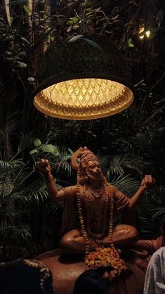 a buddha statue sitting under a chandelier surrounded by greenery