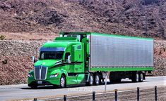 a green semi truck driving down the road in front of a mountain side with mountains behind it