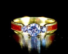 a yellow gold ring with two blue and red stones on the side, sitting on a reflective surface