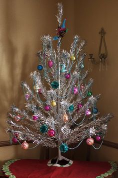 a small silver christmas tree with ornaments on it's base and red blanket underneath