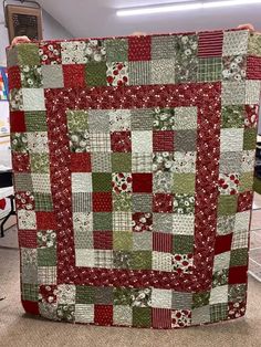 a person holding up a red and green patchwork quilt in the middle of a room