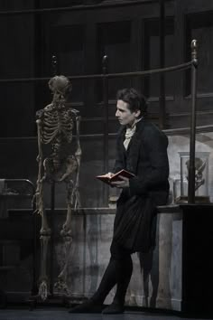 a man standing in front of a skeleton holding a book and looking at the viewer