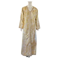 Moroccan vintage exotic metallic white gold brocade caftan gown, circa 1970s. The luxurious Moorish kaftan is designed with white and gold metallic brocade. The front of the elegant caftan gown is embellished at the front with woven gold buttons and loops that run down the center to the hemline. The cuffs and border seams are accented with handmade applique gold trim. This exotic Arabic maxi dress caftan gown makes a very extravagant garment for formal attire transitions effortlessly to casual w Gold Floor-length Kaftan For Festive Occasions, Festive Floor-length Gold Kaftan, Elegant Gold Floor-length Thobe, Festive Gold Floor-length Kaftan, Traditional Gold Floor-length Kaftan, Festive Gold Kaftan For Festive Occasions, Gold Thobe For Festive Evening Occasions, Traditional Gold Floor-length Thobe, Traditional Gold Floor-length Abaya