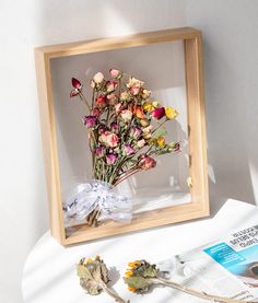 dried flowers in a wooden frame on top of a table next to a news paper