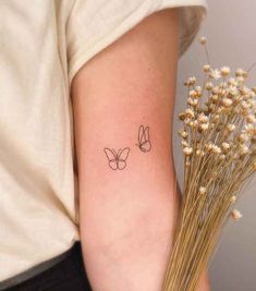 a small butterfly tattoo on the left arm and behind it is a plant with white flowers