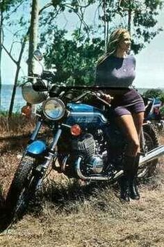 a woman sitting on top of a motorcycle in the grass next to trees and water