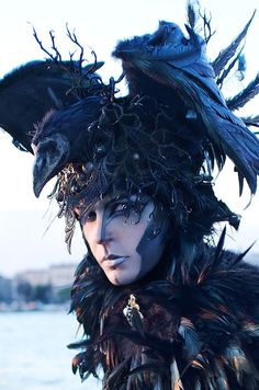 a woman with black feathers on her head and face is standing in front of the water